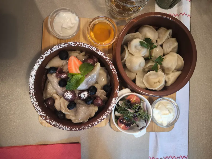 Wareniki mit Kirschen und Beeren neben Pelmeni im Tontopf, auf einem bestickten Tischläufer